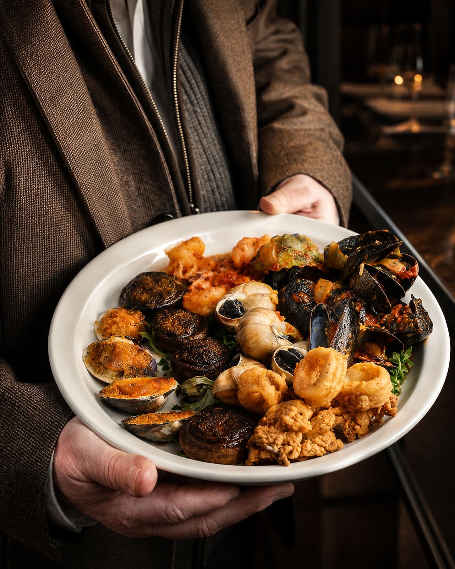 A server carrying a large white platter of baked stuffed clams, stuffed mushrooms, escargots in the shell, mussels in red sauce and fried calamari through a dark dining room.