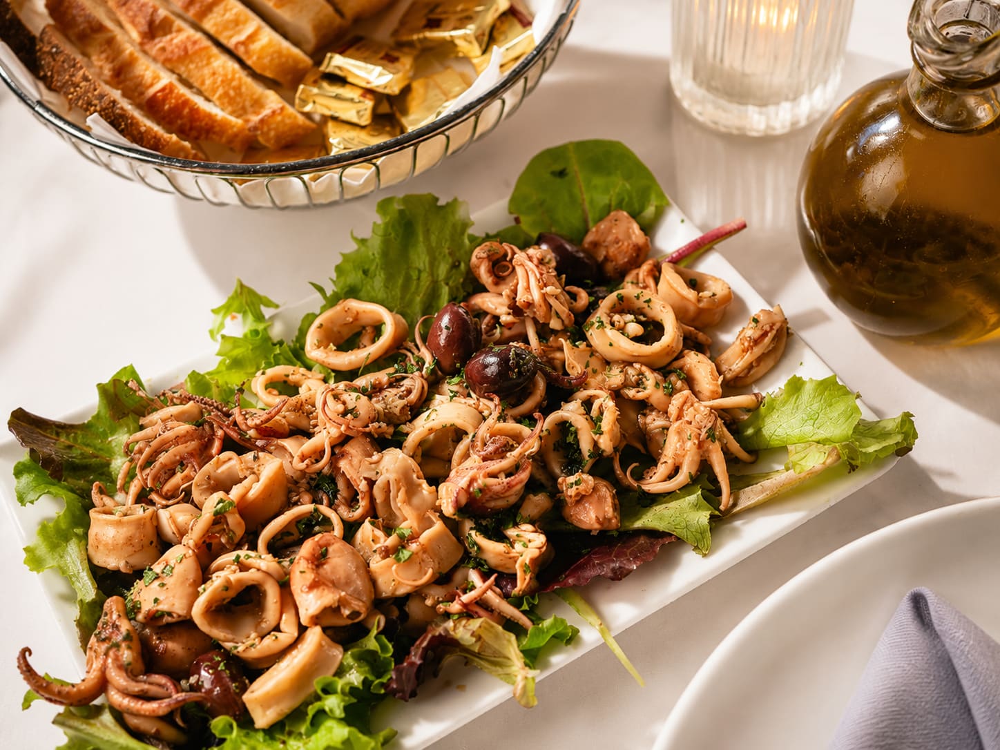 A long white plate of calamari rings and octopus over lettuce with black olives, beside a bread basket and a cruet of olive oil.