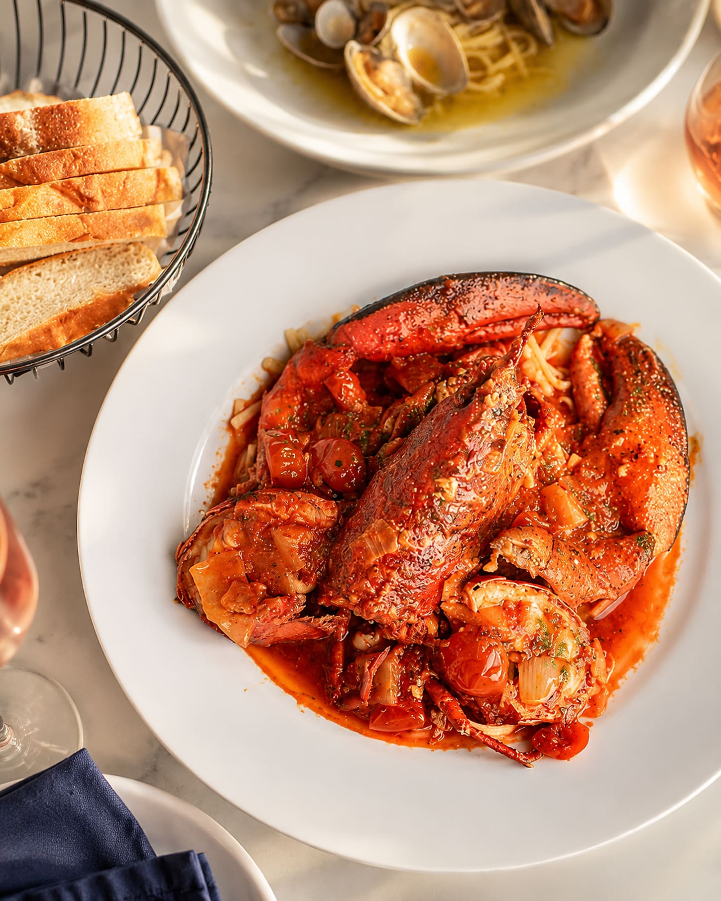 A lobster in spicy red sauce over linguine on a white plate, claws and tail shell in the sauce, with a bread basket and a glass of rosé alongside.