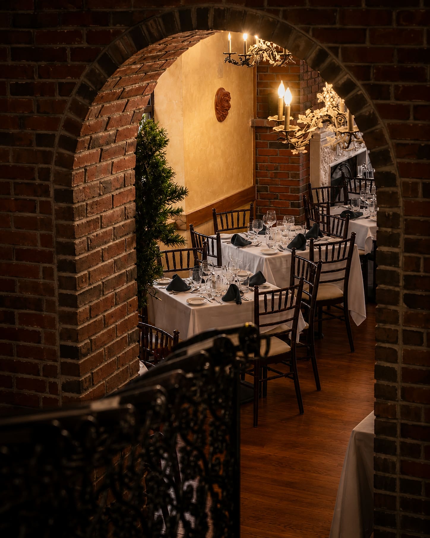White-clothed tables set with wine glasses and folded napkins, seen through a brick archway under a chandelier.