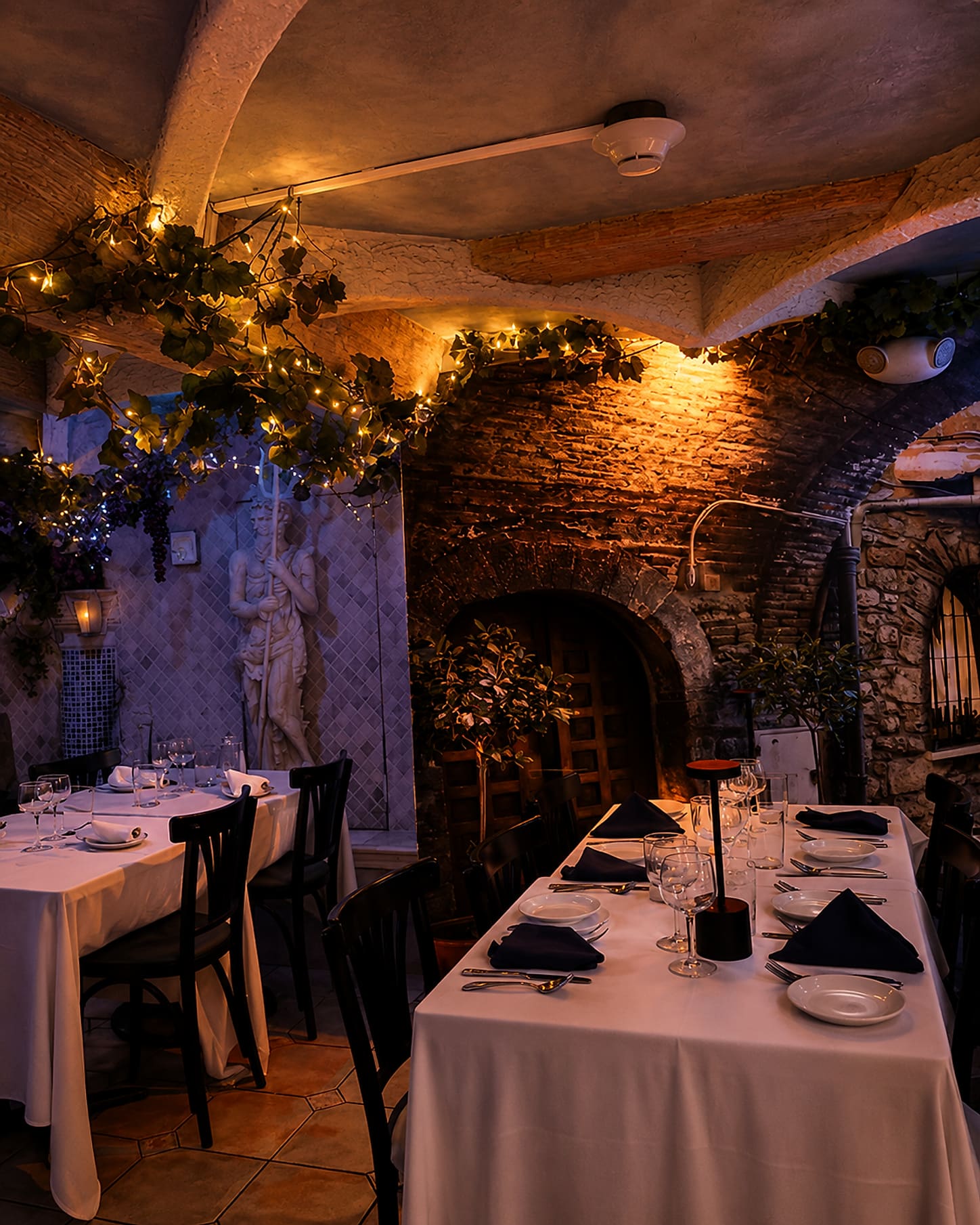 A stone-arched cellar dining room lit warmly, with white-clothed tables, dark chairs and greenery trained across the vaulting.
