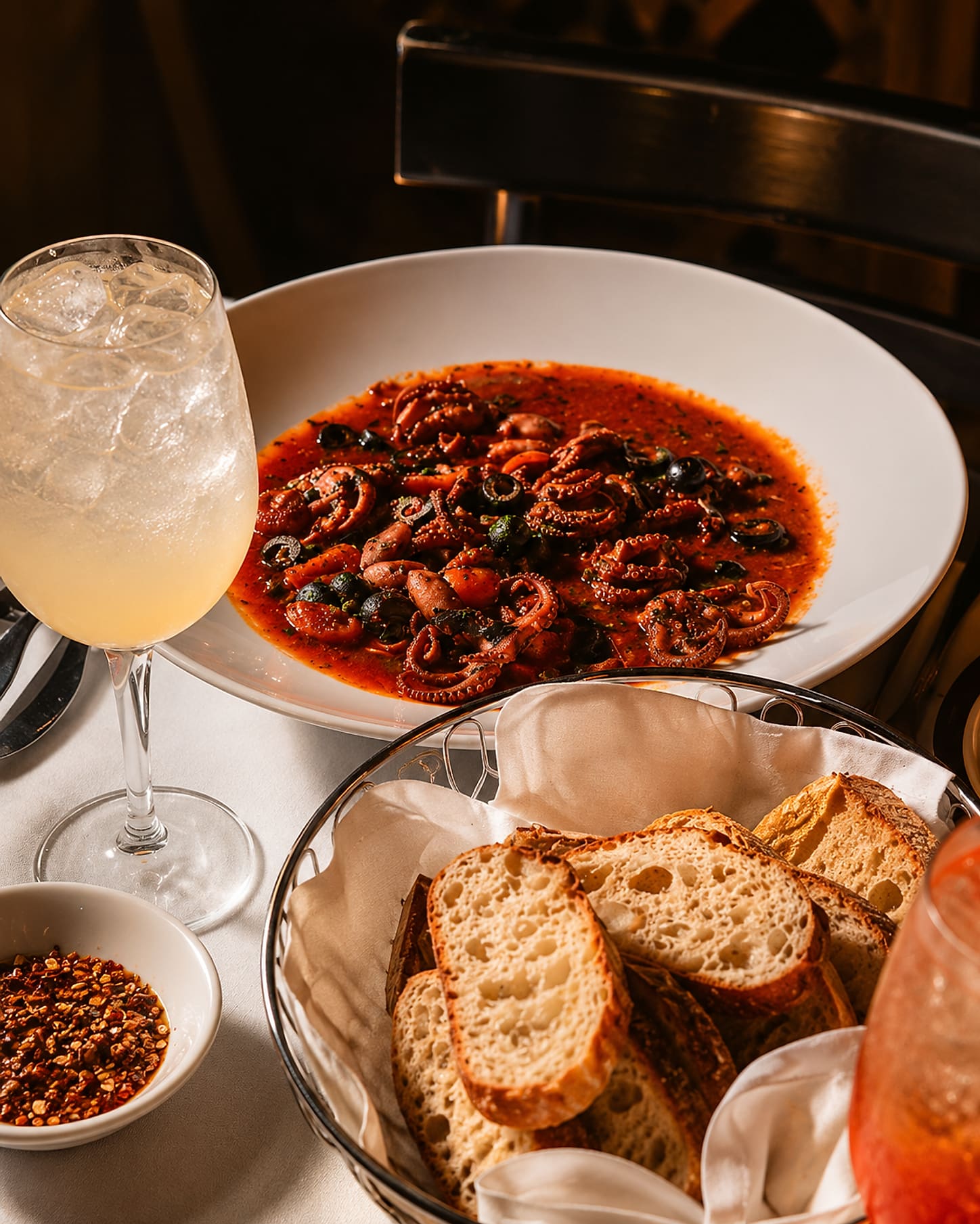 A wide white bowl of baby octopus in red sauce with black olives and cherry tomatoes, beside a basket of sliced bread and a dish of crushed red pepper.