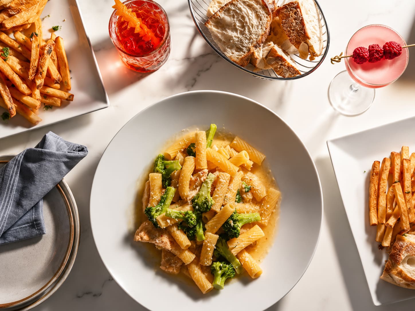 Rigatoni with broccoli in a light sauce in a white bowl, shot from above on marble with bread, two cocktails and plates of chips around it.