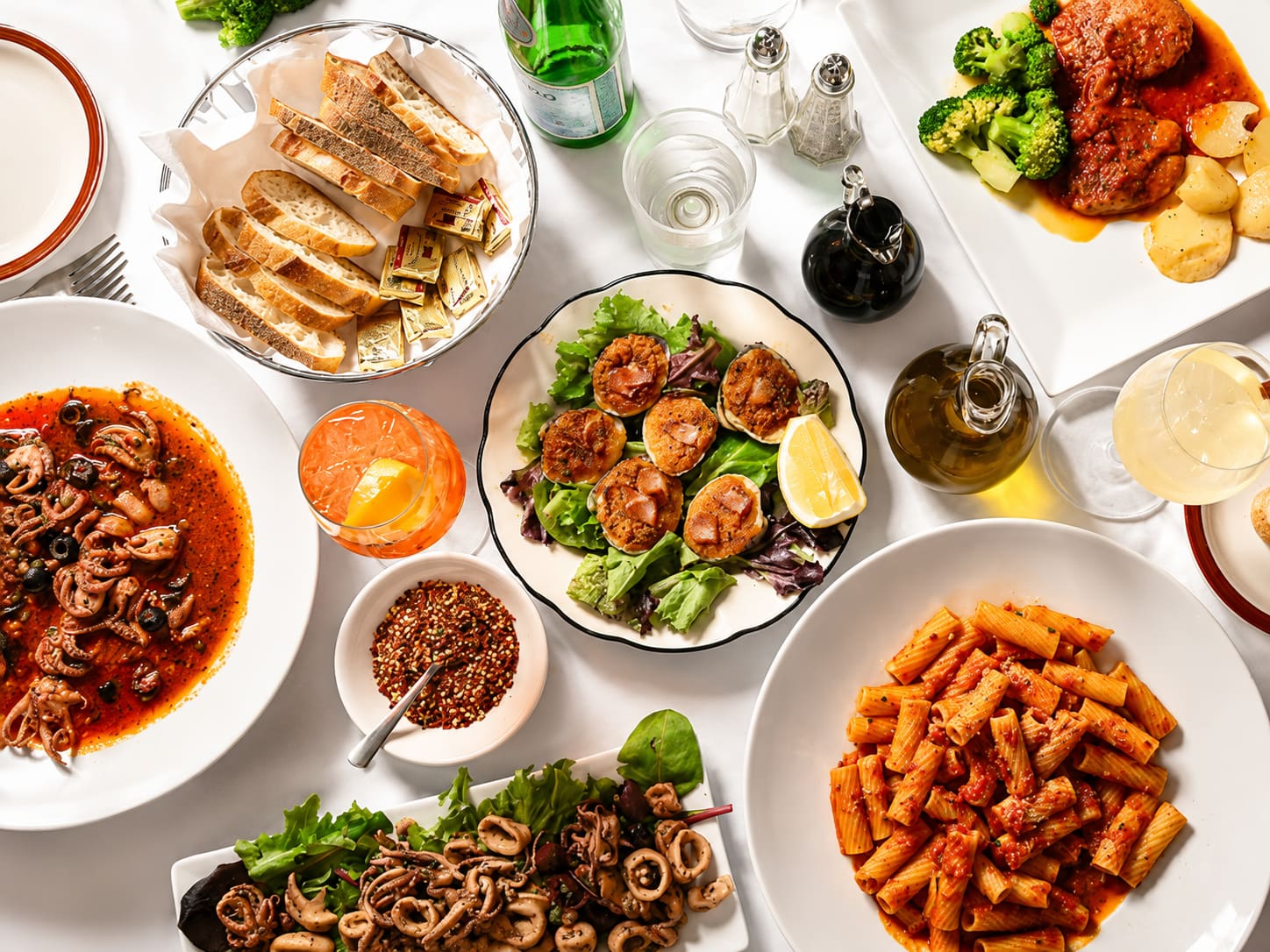 A table photographed from above, crowded with a bowl of octopus in red sauce, a calamari salad, rigatoni in tomato sauce, sliced bread, olive oil and a dish of crushed red pepper.