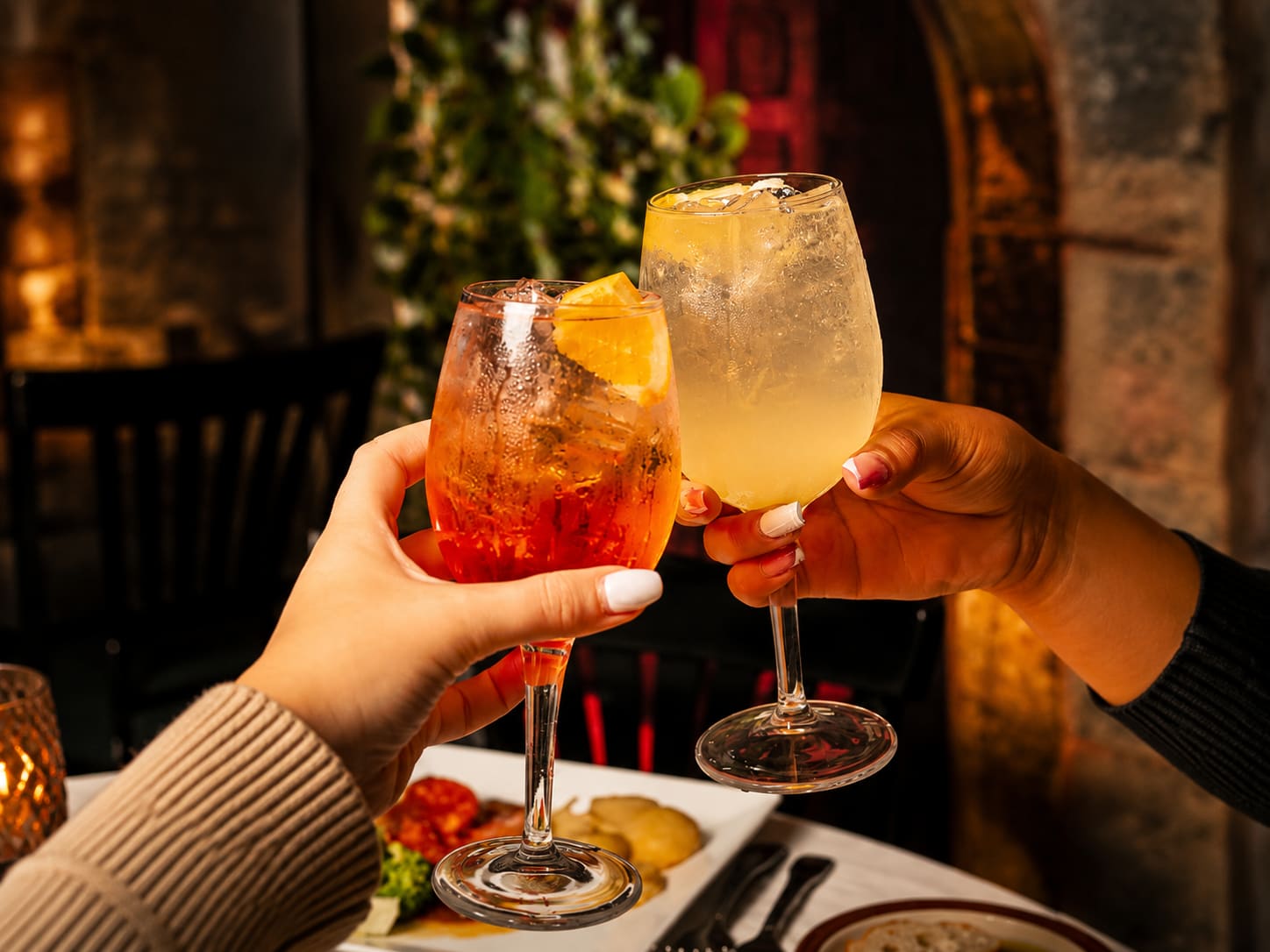 Two hands touching glasses together over a table — a spritz and a tall citrus cocktail — in a warm, dark, stone-walled room.